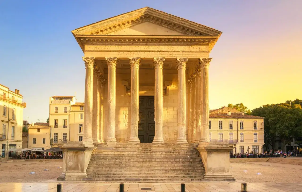 Maison Carr&eacute;e, a Roman temple in N&icirc;mes, France, bathed in golden sunset light. People dine at outdoor cafes in the plaza.