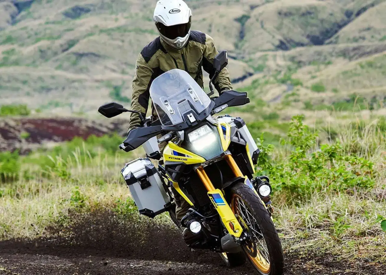 Motociclista en una maxi trail amarilla y blanca, con alforjas, recorriendo un camino de tierra con vegetaci&oacute;n y colinas al fondo.
