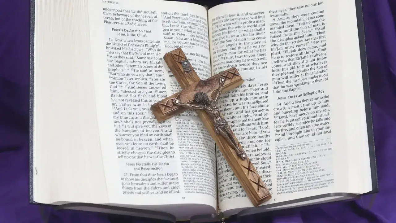 A wooden crucifix rests on an open Bible, with text from Matthew 16 visible, hinting at homilies for the 33rd Sunday of Year B.