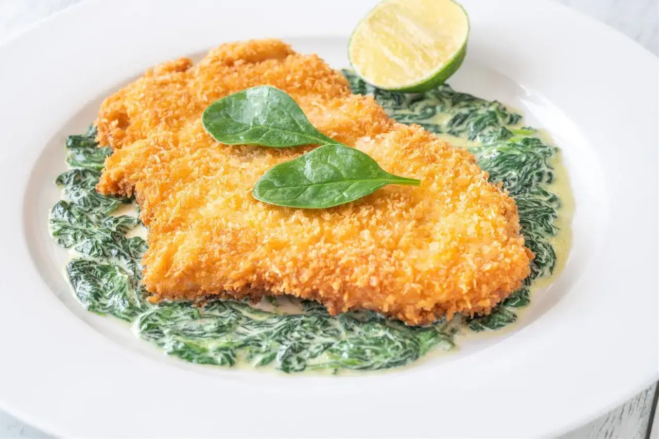Chrupiący kotlet milanese na kremowym szpinaku, udekorowany listkami szpinaku i połówką limonki.