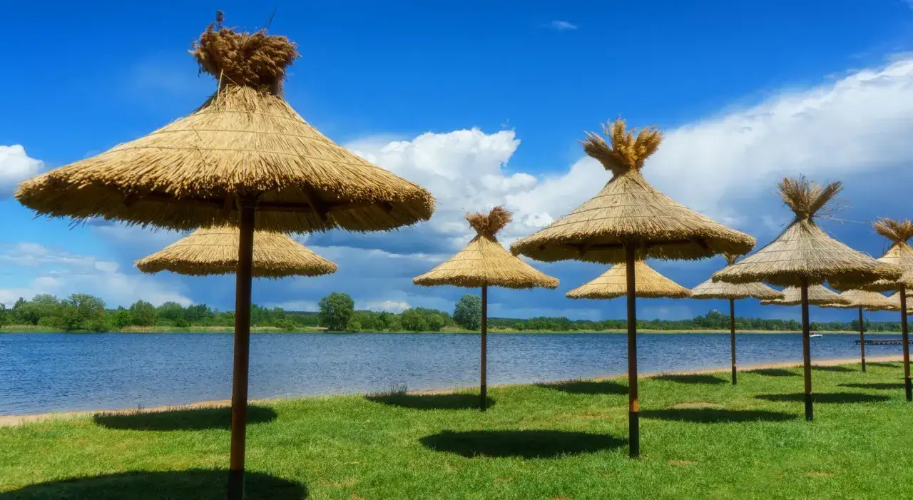 Słomiane parasole nad brzegiem rzeki, zapraszają do odpoczynku. Błękitne niebo z chmurami i zieleń trawy tworzą idylliczny krajobraz.