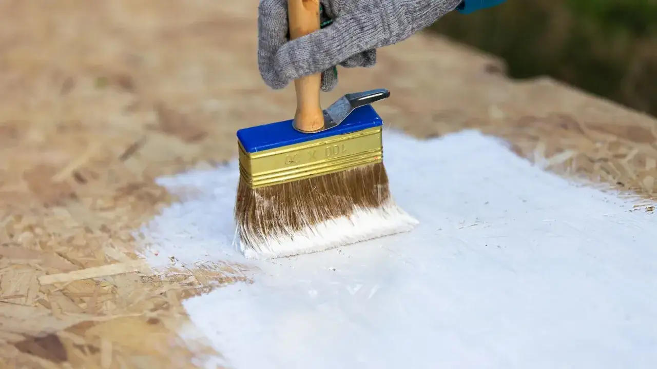 En hand med handske målar OSB-skiva vit med en bred pensel.