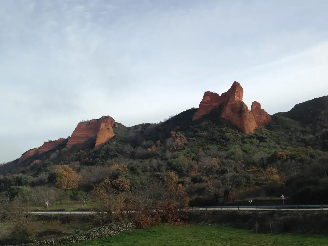 Majestuosas formaciones rojas se alzan sobre un paisaje verde y otoñal. Un camino serpentea al pie de las montañas, cerca de un merendero junto al lago de Carucedo.