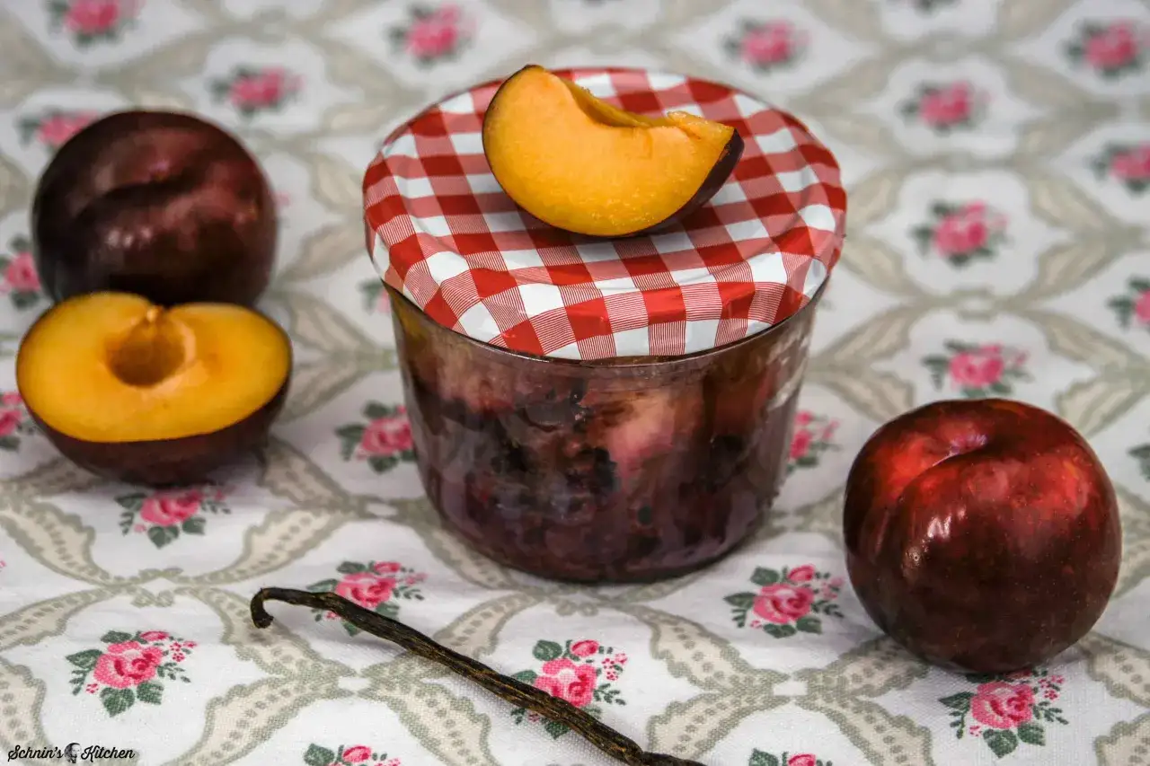 Ein Glas mit Pflaumenkompott, frische Pflaumen und eine Vanilleschote. So kann man Pflaumen aus dem Glas lecker verarbeiten.