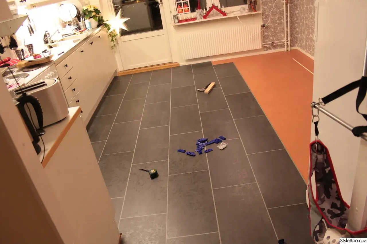 Ett k&ouml;ksgolv med m&ouml;rkgr&aring; klinkerplattor som h&aring;ller p&aring; att l&auml;ggas. Verktyg och bl&aring; plastbitar ligger utspridda, redo att m&aring;la linoleumgolv.