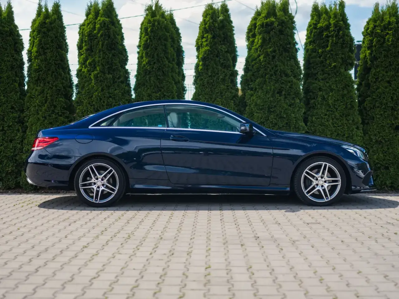 Elegancki, granatowy w212 coupe stoi na wybrukowanym podjeździe, otoczony rzędem wysokich, zielonych żywotników.