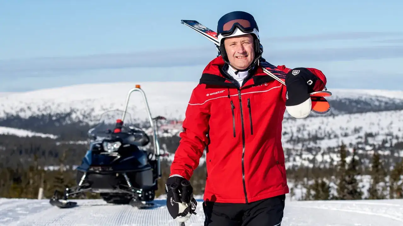 Skistar VD med skidor på axeln och snöskoter i bakgrunden.