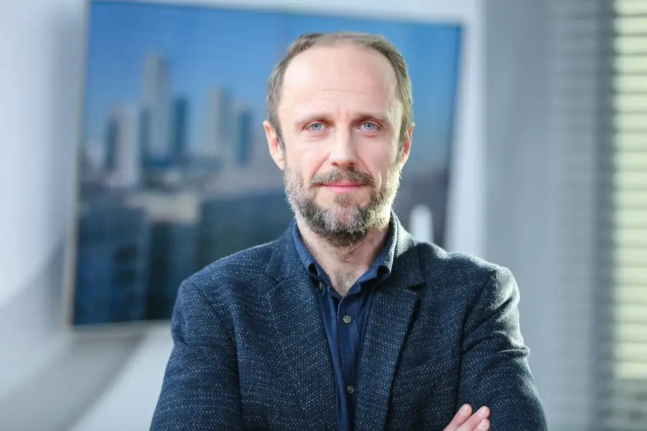 Łukasz Simlat, znany z wielu filmów, w eleganckim garniturze, z niebieskimi oczami i zarostem.