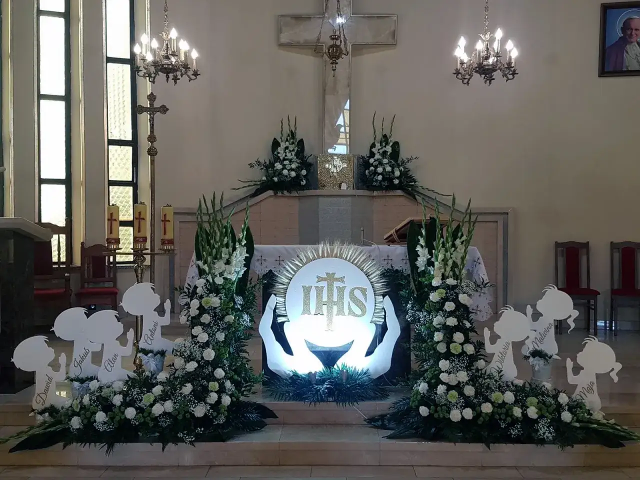 Ołtarz w kościele, ozdobiony kwiatami i symbolami religijnymi. Centralnie umieszczony symbol IHS, otoczony dłońmi. Obok dziecięce sylwetki. To nie jest świętokradztwo, lecz piękna dekoracja.