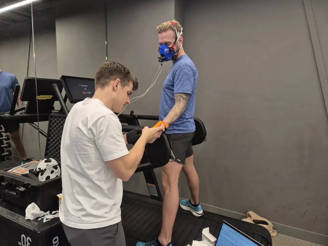 Un athlète porte un masque respiratoire sur un tapis roulant, tandis qu'un entraîneur ajuste un appareil. Le test vise à mesurer les performances et l'accumulation d'acide lactique.