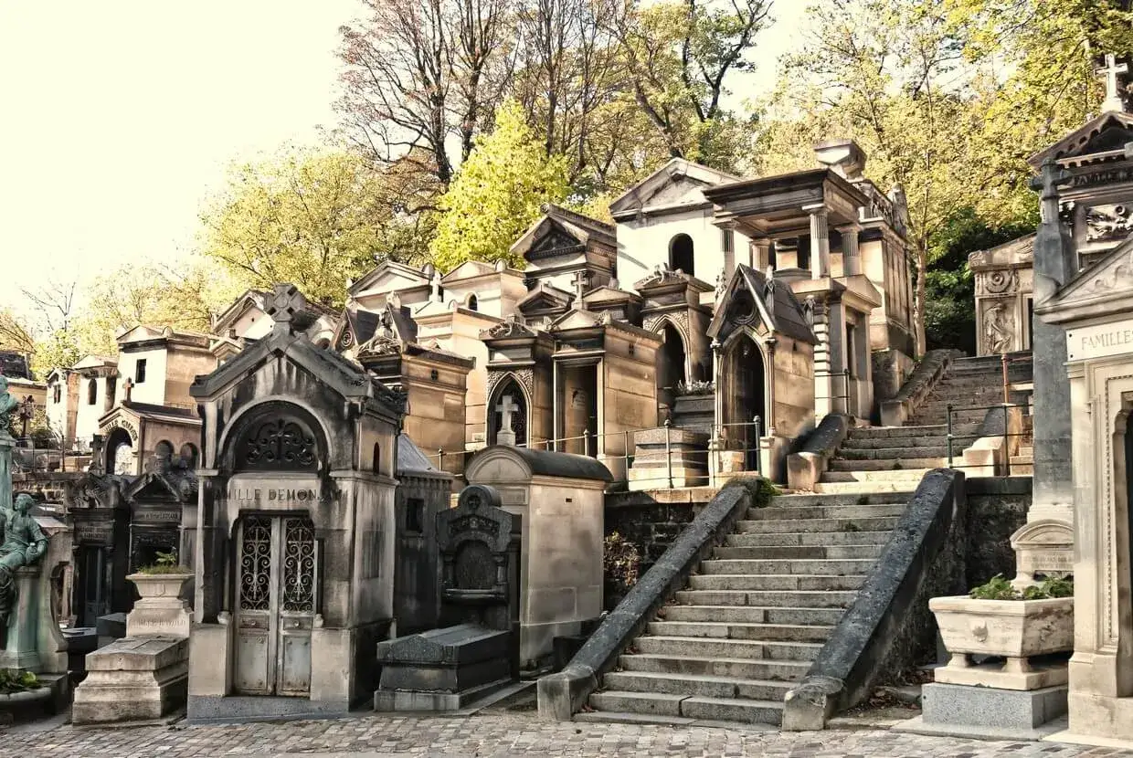 Kamienne grobowce i schody na cmentarzu Père Lachaise, Paryż. Wiosenne drzewa tworzą tło dla architektonicznych detali.
