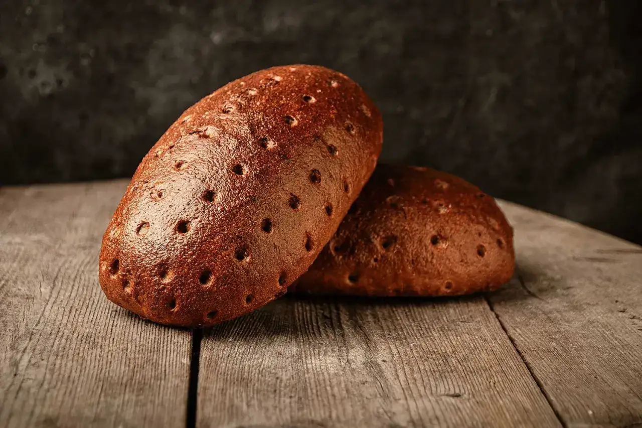 Zwei dunkle, rustikale Brote mit vielen kleinen Löchern liegen auf einer Holzplatte. Perfekt für das Brot backen.
