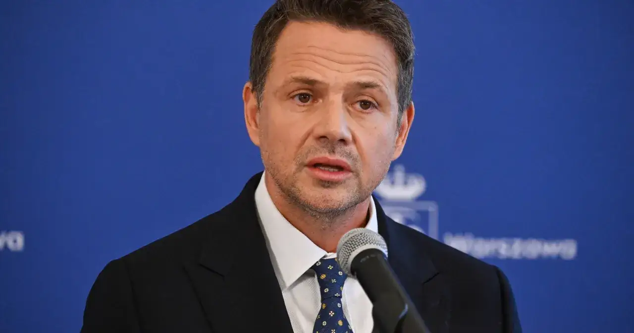 Prezydent Warszawy w garniturze i krawacie przemawia do mikrofonu na tle niebieskiej ściany z logo miasta.