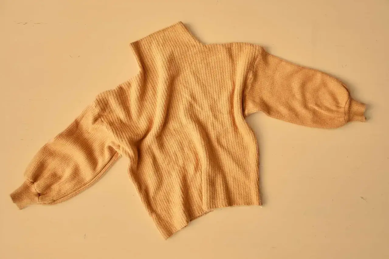 Sfilcowany sweter w kolorze musztardowym, z bufiastymi rękawami i golfem, leży na jasnym tle.