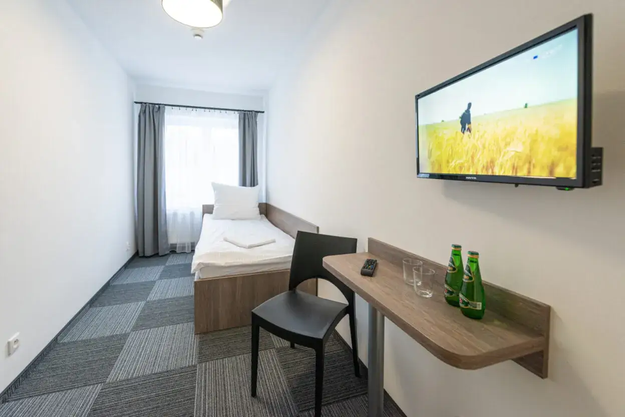 Pok&oacute;j w hostelu: ł&oacute;żko, krzesło, stolik z napojami i pilotem, telewizor. To idealne miejsce na kr&oacute;tki pobyt.