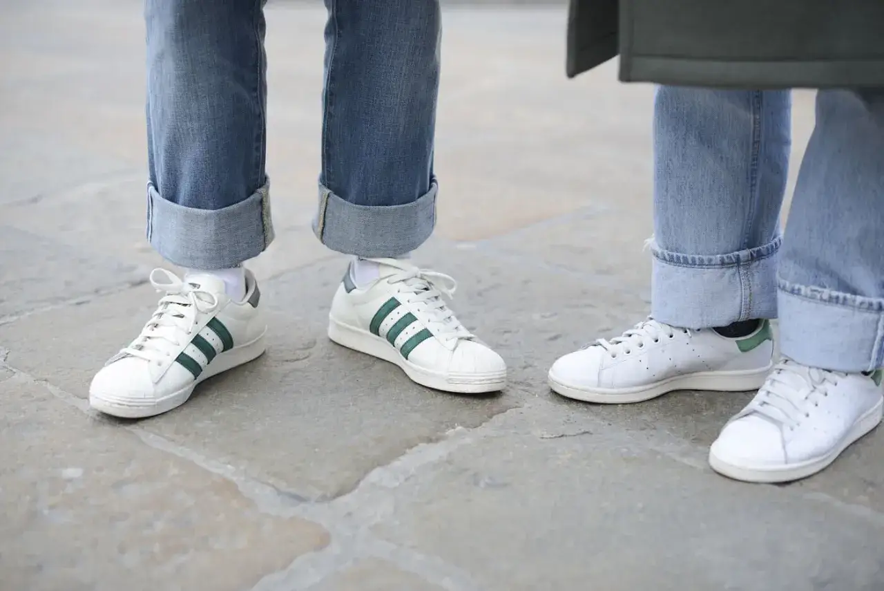 Dwie pary białych adidasów z zielonymi paskami i zielonymi elementami na pięcie, zestawione z podwiniętymi jeansami. Czy adidas ma zawyżoną rozmiarówkę?