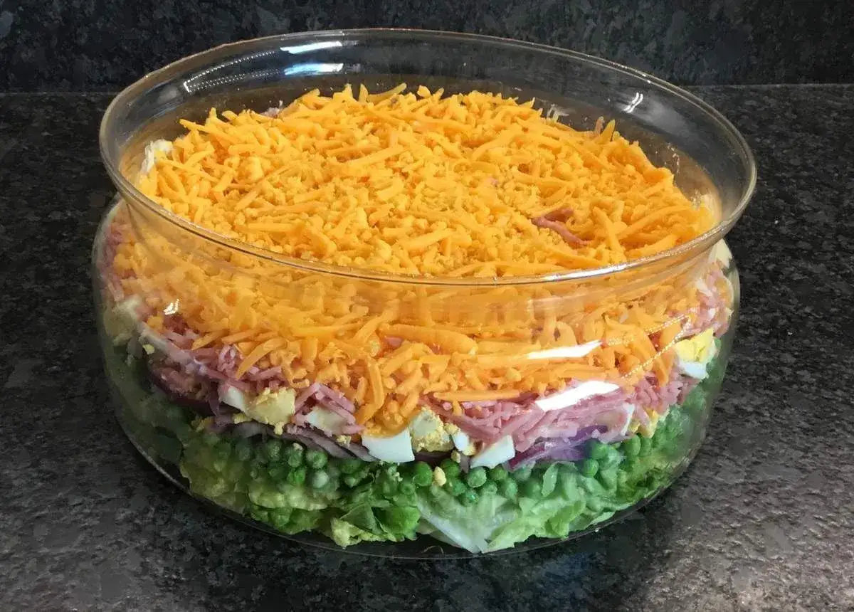 Une salade 7 &eacute;tages color&eacute;e dans un grand bol en verre, avec des couches de laitue, petits pois, oignons rouges, jambon, &oelig;ufs durs et fromage r&acirc;p&eacute;.