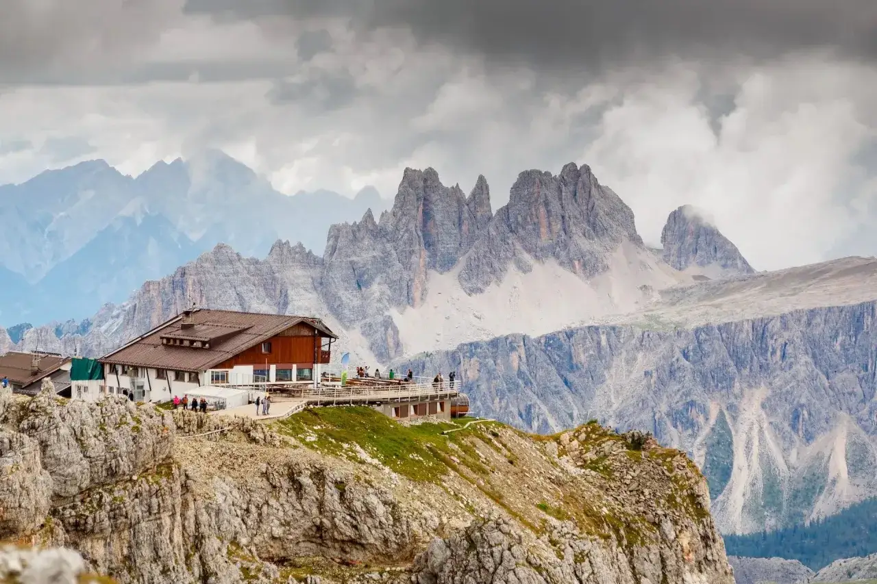 Schronisko na szczycie, punkt startowy Alta Via 1, z widokiem na skaliste szczyty Dolomitów pod pochmurnym niebem.