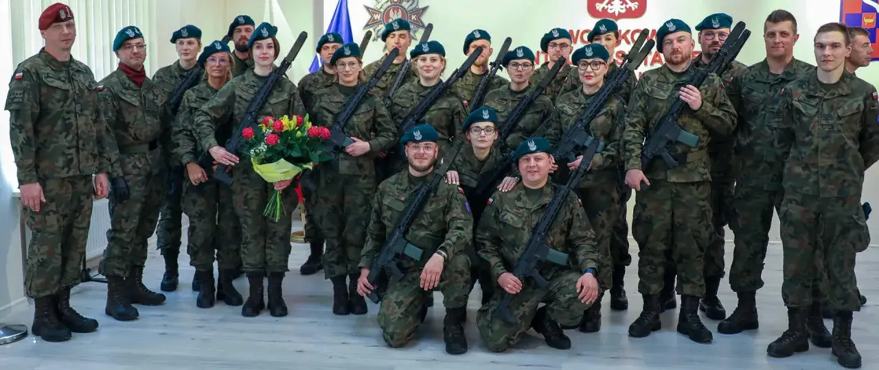 Grupa żołnierzy po szkoleniu wojskowym pozuje do zdjęcia z bronią i bukietem kwiatów.