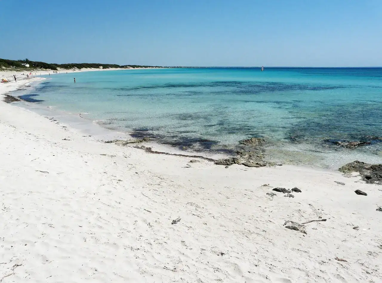 Spiaggia puglia con sabbia bianca e mare cristallino. In lontananza, una persona fa windsurf.