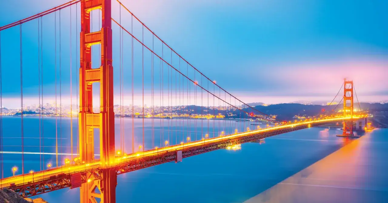 Złota Brama, **most w San Francisco**, lśni w blasku świateł o zmierzchu, rozświetlając wodę i miasto w oddali.