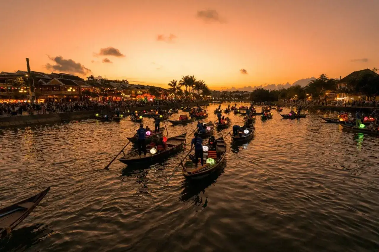 Boote mit Laternen auf einem Fluss vor der beleuchteten Hoi an Altstadt bei Sonnenuntergang.