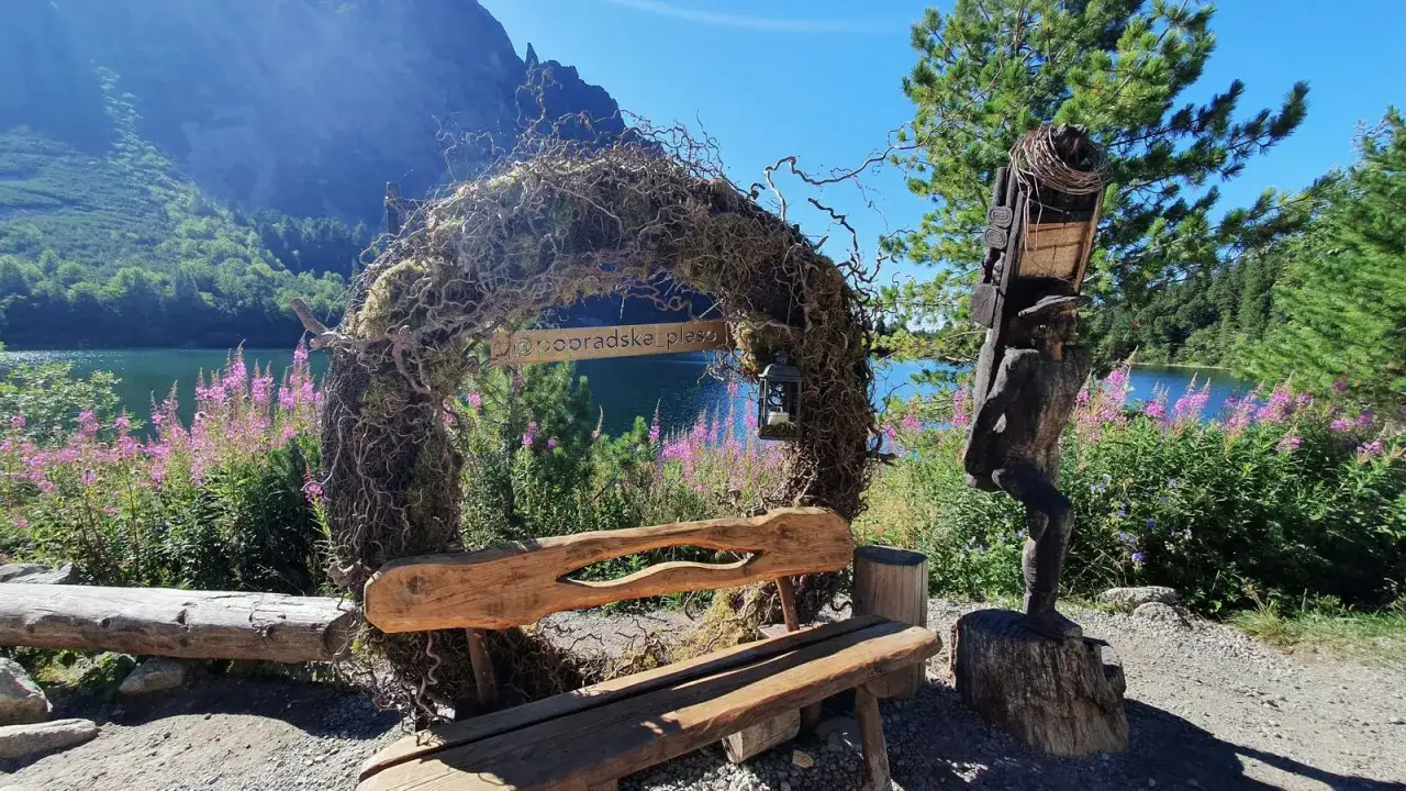 Drewniana rzeźba i ławka przy wejściu na parking Popradzkie Pleso. W tle jezioro i góry.