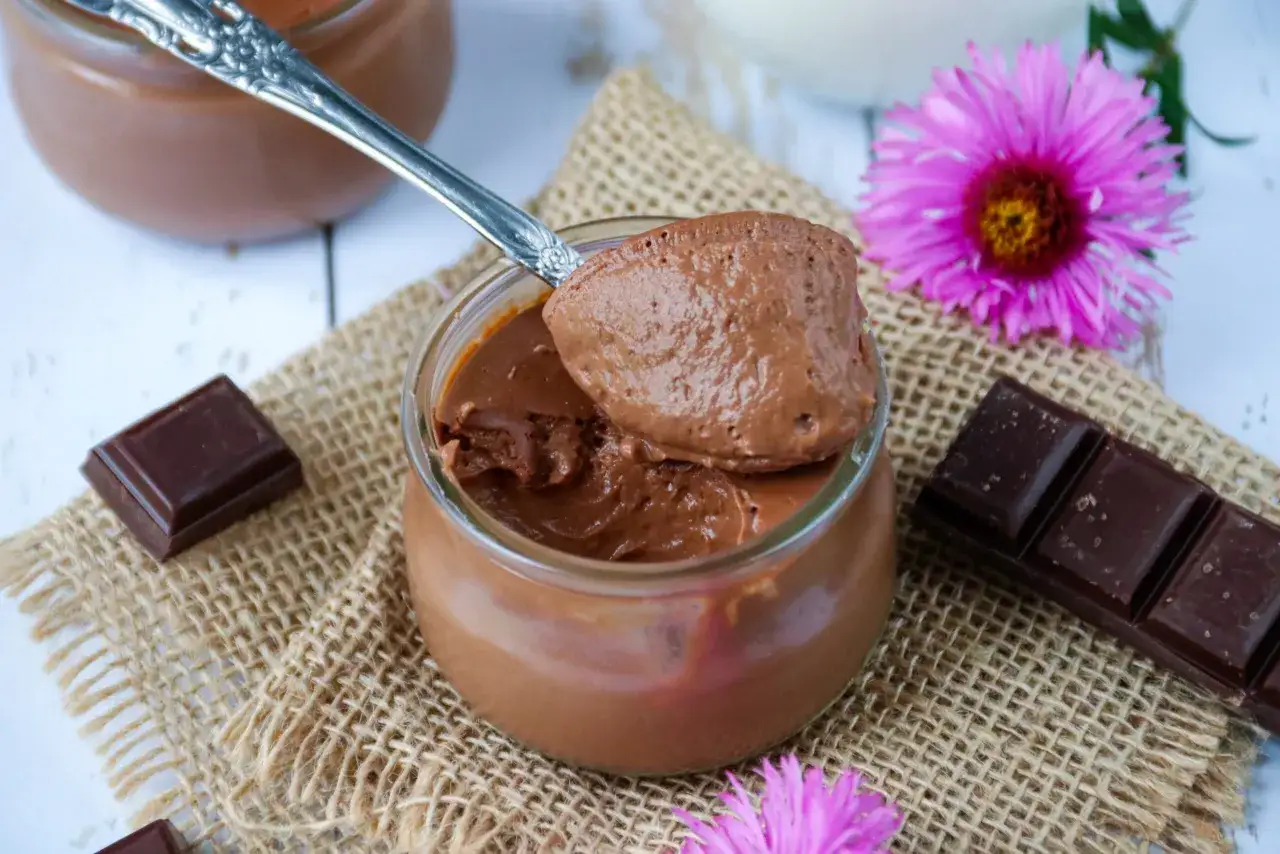 Une cuillère de crème au chocolat sans sucre, onctueuse et aérienne, sur un pot en verre. Des carrés de chocolat et des fleurs roses décorent la scène.