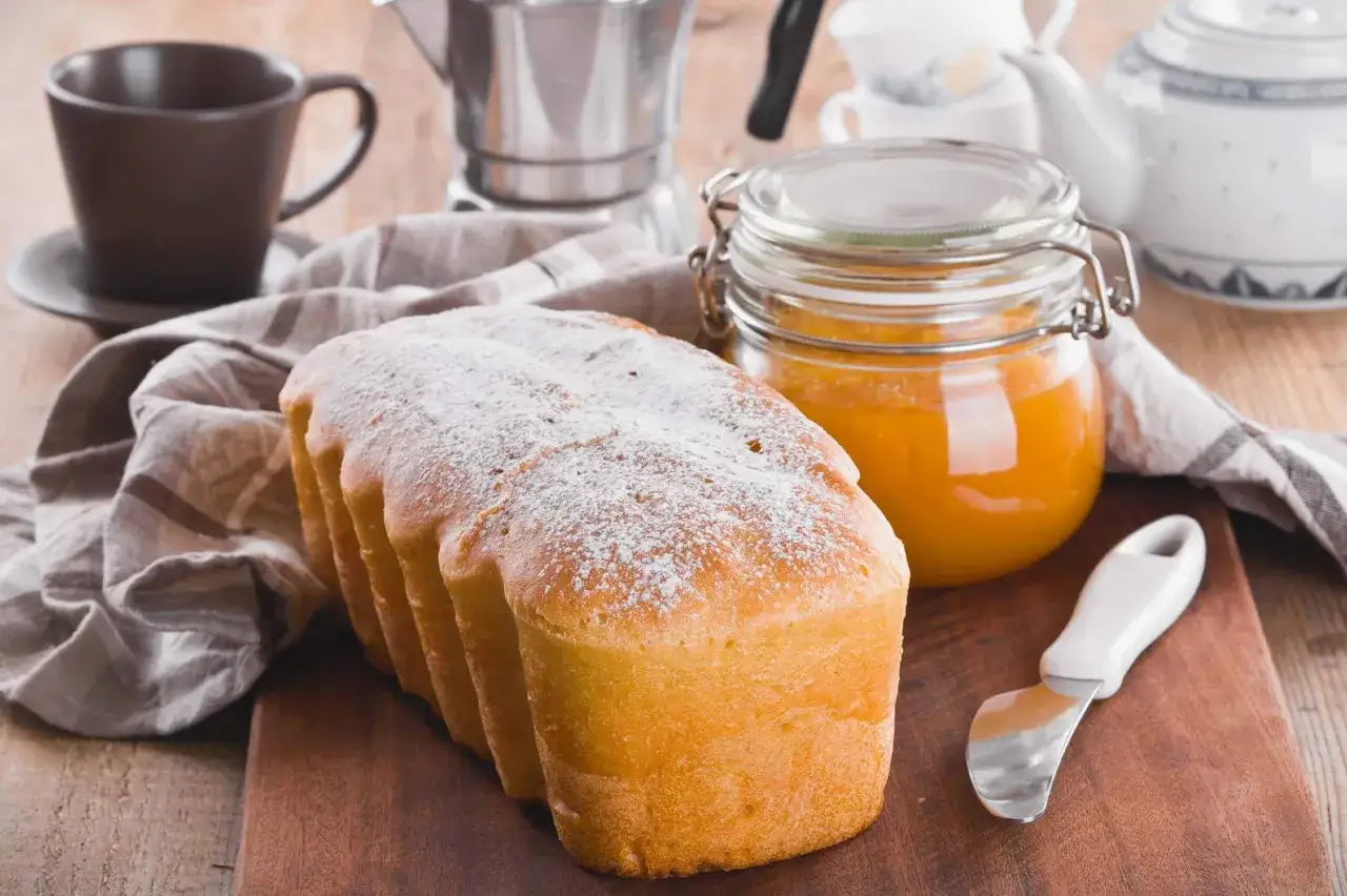 Plumcake all'arancia soffice, spolverato di zucchero a velo, accanto a un barattolo di marmellata d'arancia e una tazzina di caffè.