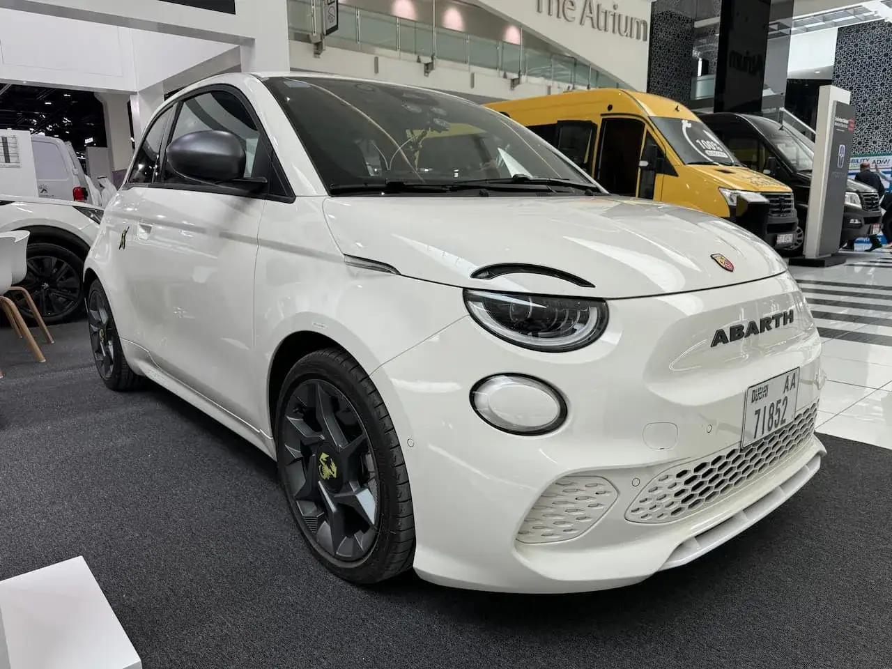 Biały Abarth 500e z czarnymi felgami i logo skorpiona. Auto prezentowane w salonie, w tle żółty van.