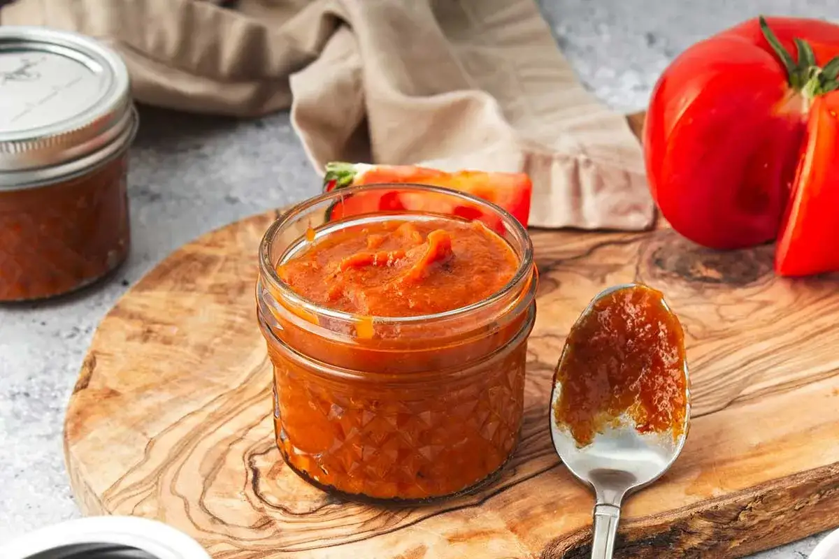 Ein Glas mit selbstgemachtem, veganem Ketchup steht auf einem Holzbrett. Frische Tomaten liegen daneben.