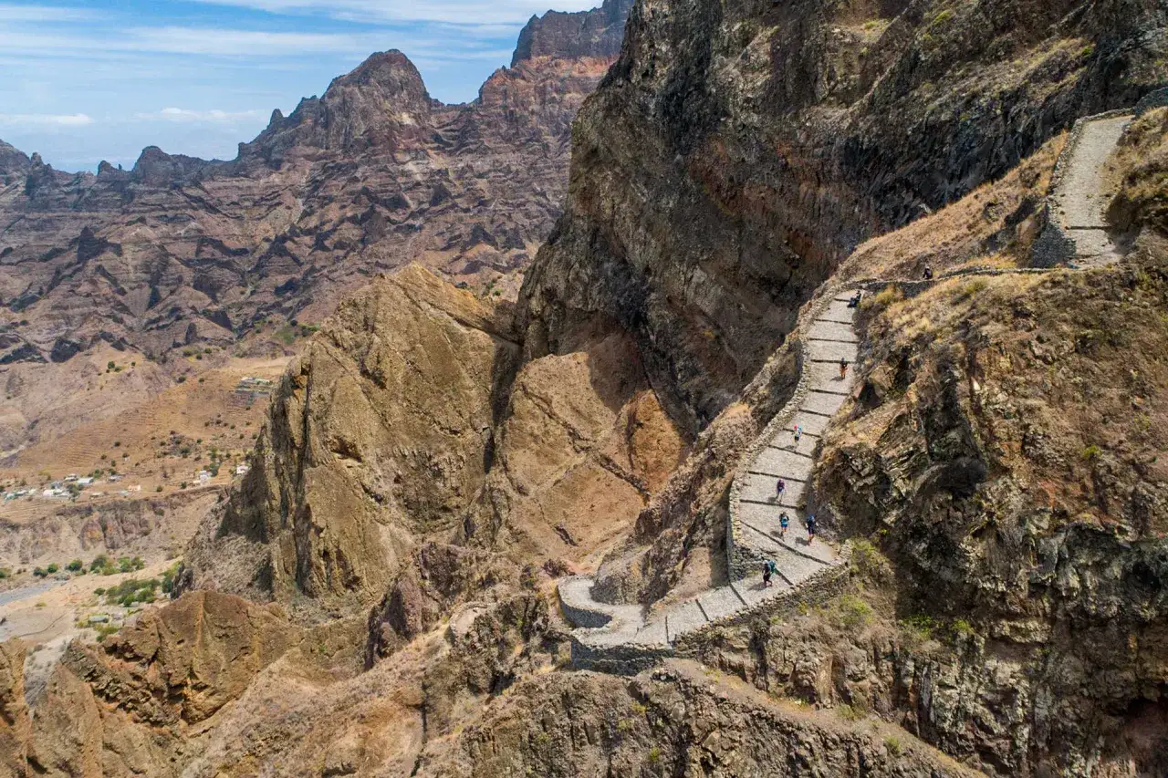 Une **randonnée** spectaculaire sur un sentier en pierre à flanc de montagne, avec des randonneurs profitant du paysage aride du **Cap Vert**.