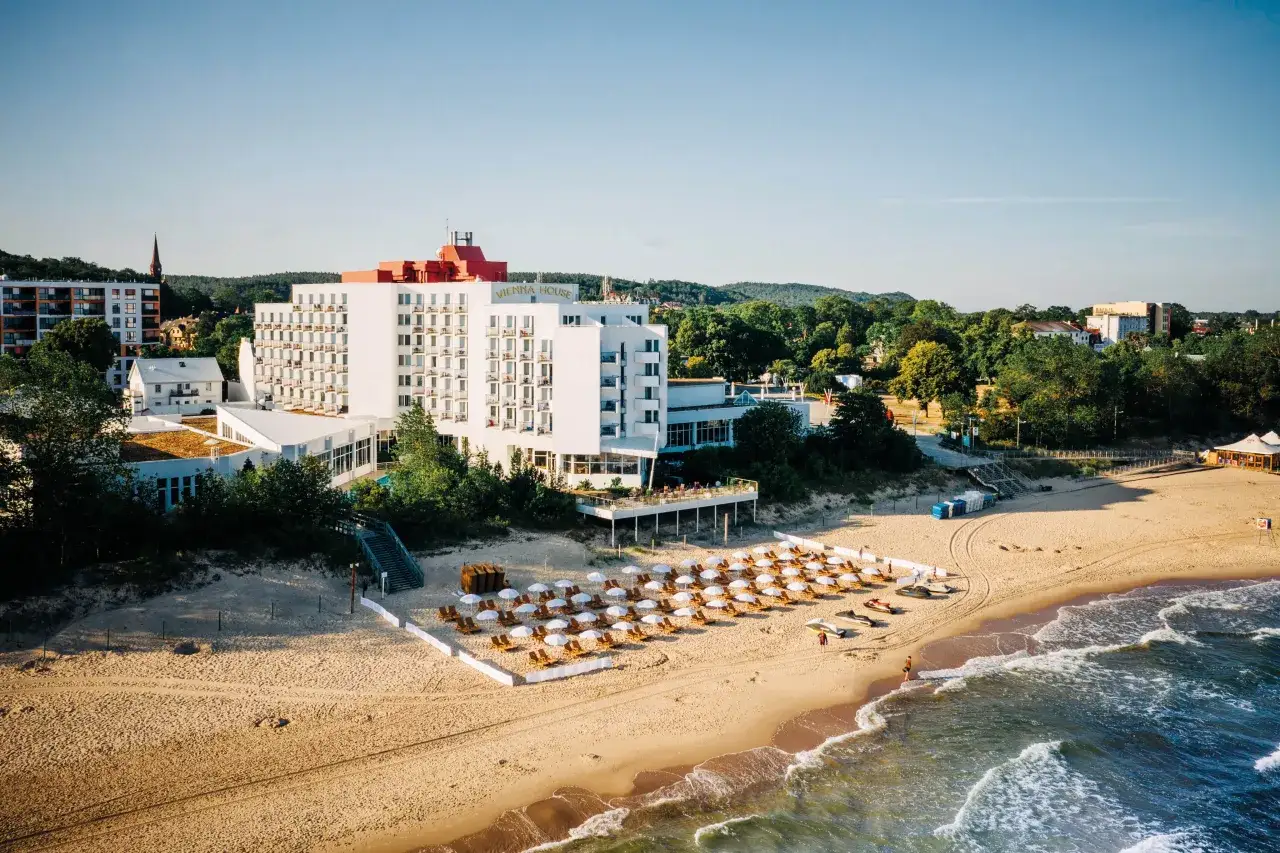 Ośrodek wczasowy Amber wita gości na piaszczystej plaży. Biały budynek hotelu z czerwonym dachem góruje nad leżakami i parasolami.