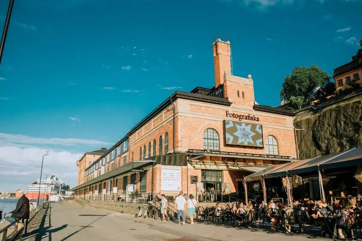 Solig dag vid Fotografiska Stockholm. M&auml;nniskor sitter vid uteserveringen framf&ouml;r den tegelbyggda konsthallen.