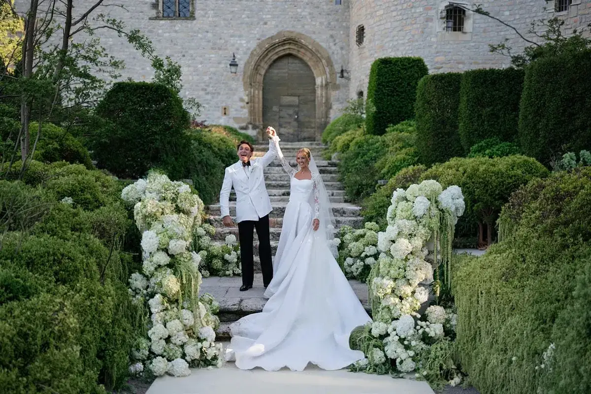 Brudparet, med en glädje som en Desiré Inglander, firar på en stenlagd trappa, omgiven av frodig grönska och vita blommor.