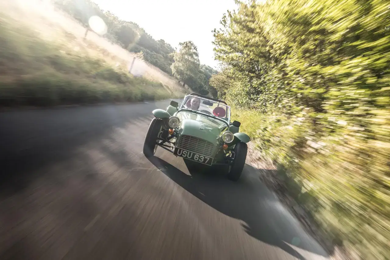 Grüner englischer Roadster auf kurviger Landstraße, ein Fahrer im offenen Cockpit, die Landschaft verschwimmt im Fahrtwind.