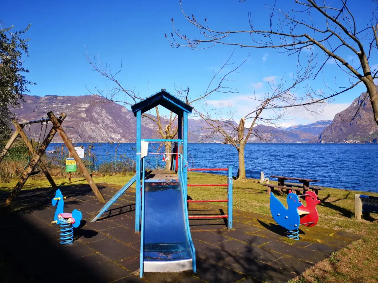 Area giochi con altalena e scivolo sul lago di Barcis spiaggia, con montagne sullo sfondo.