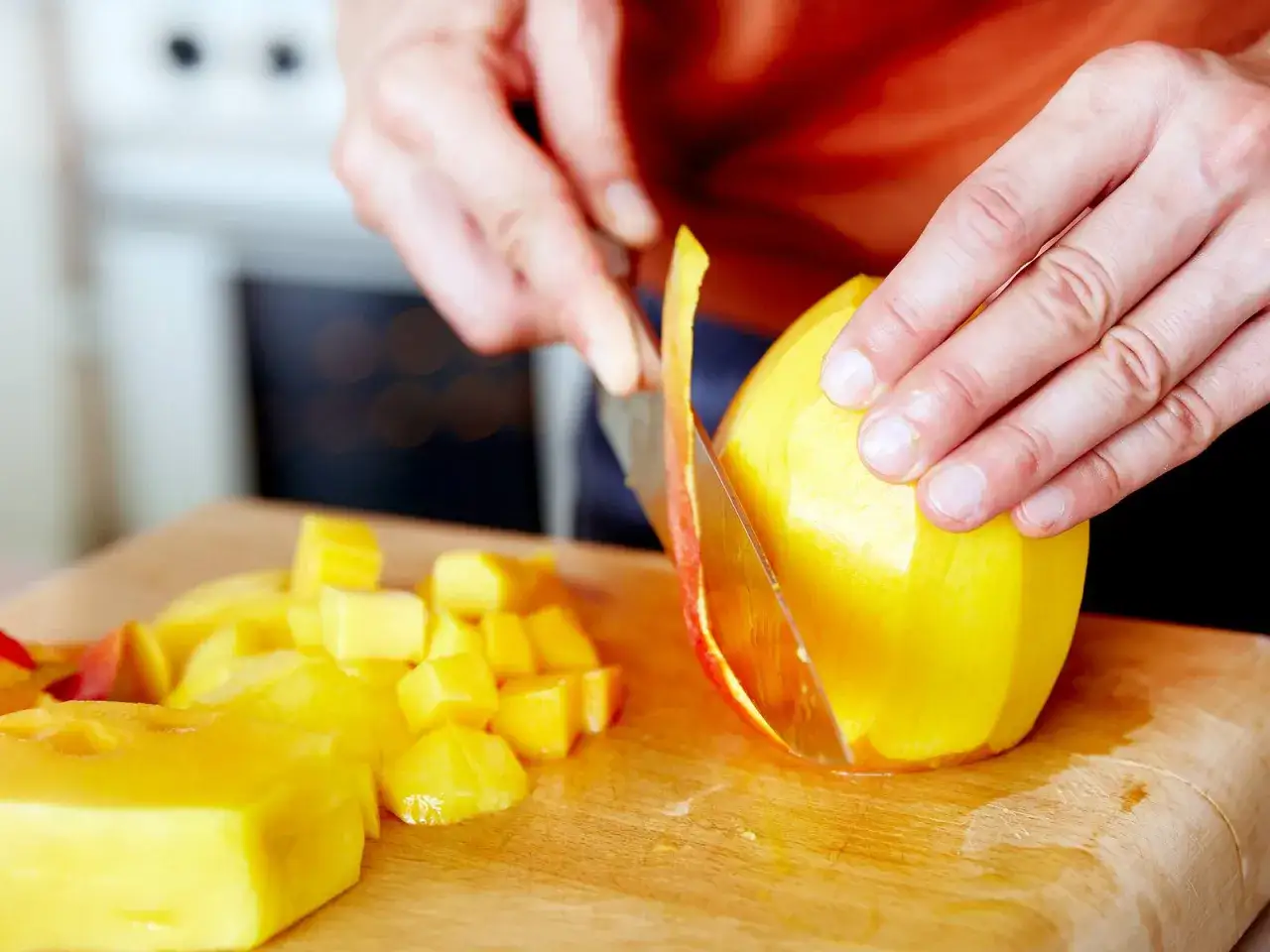 Hände schneiden eine reife Mango auf einem Holzbrett. Saftige, gelbe Würfel liegen bereit.