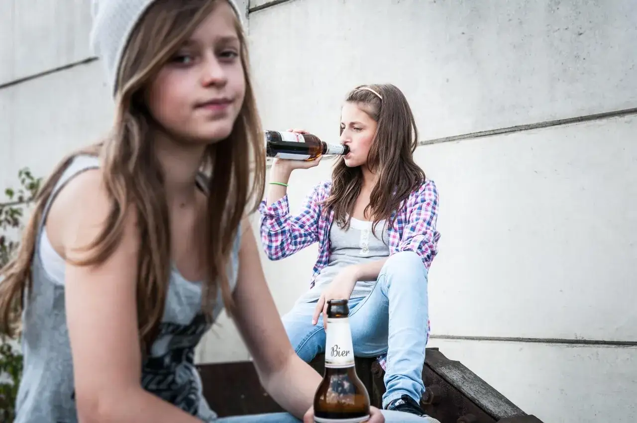 Dwie nastolatki piją piwo, co może być początkiem alkoholizmu wśród młodzieży. Jedna z nich trzyma butelkę, druga pije prosto z niej.