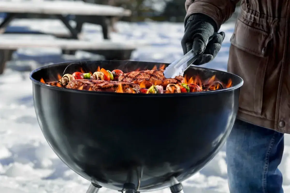 Un homme prépare un barbecue hivernal. Des brochettes de légumes et des steaks grillent sur le feu, dans un décor enneigé.