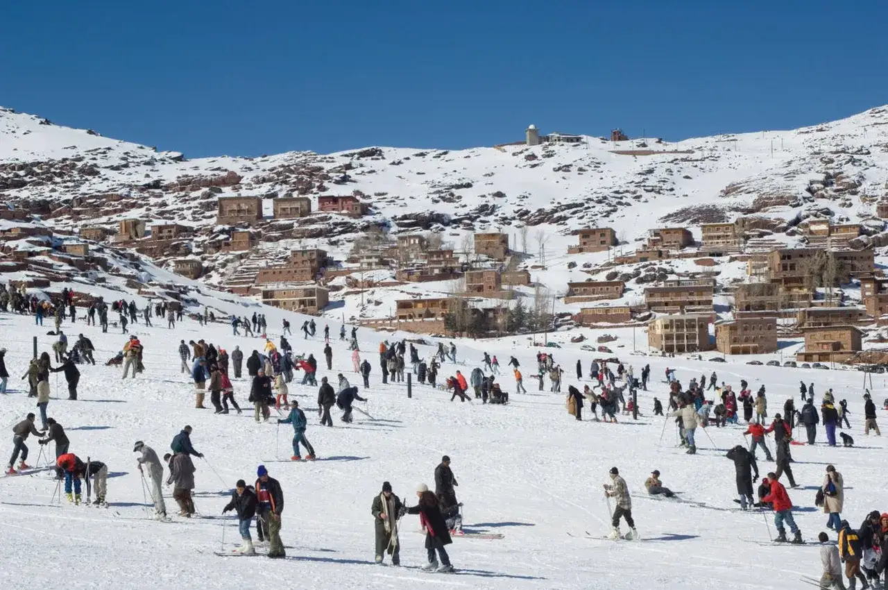 Maroko pogoda styczeń: ludzie jeżdżą na nartach i snowboardach na ośnieżonych zboczach gór Atlas, z wioską w tle.