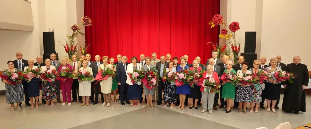 Uroczystość z okazji 60 rocznicy ślubu. Pary z bukietami kwiatów stoją na tle czerwonej kurtyny.