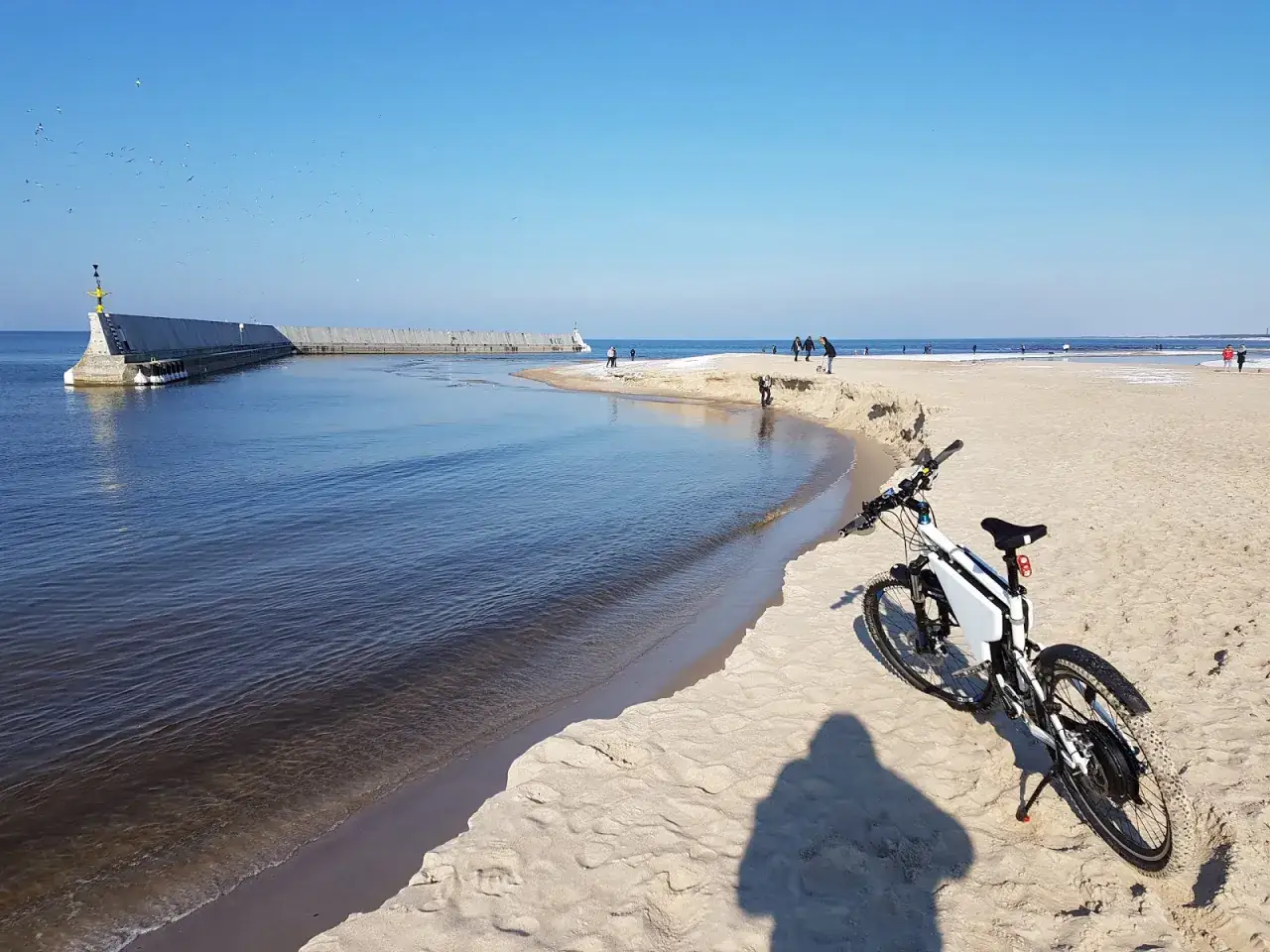 Rower stoi na piaszczystej plaży, obok spokojnej wody. W oddali widać falochron i spacerujących ludzi.