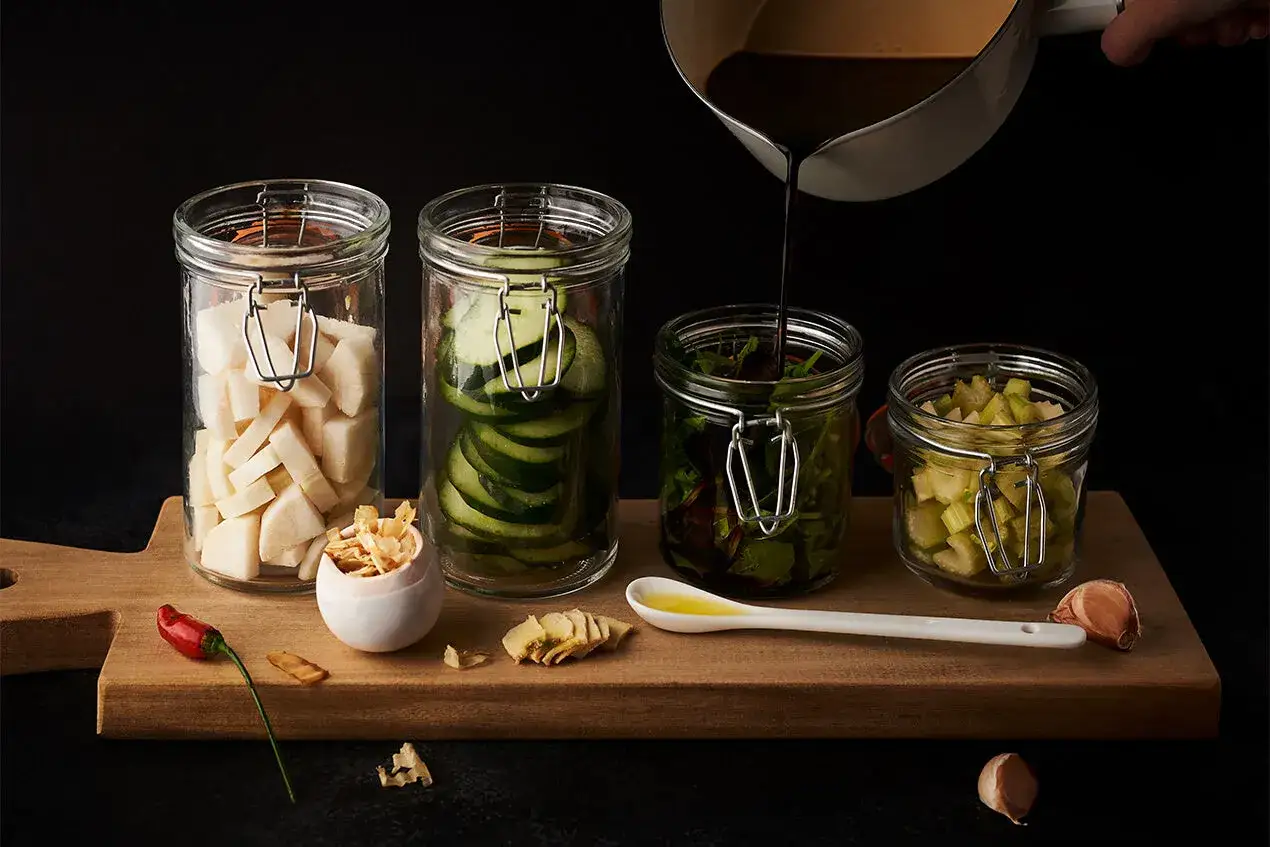 Preparing a fukujinzuke recipe: jars of daikon, cucumber, greens, and celery are ready for pickling. A dark liquid is poured from a pot.