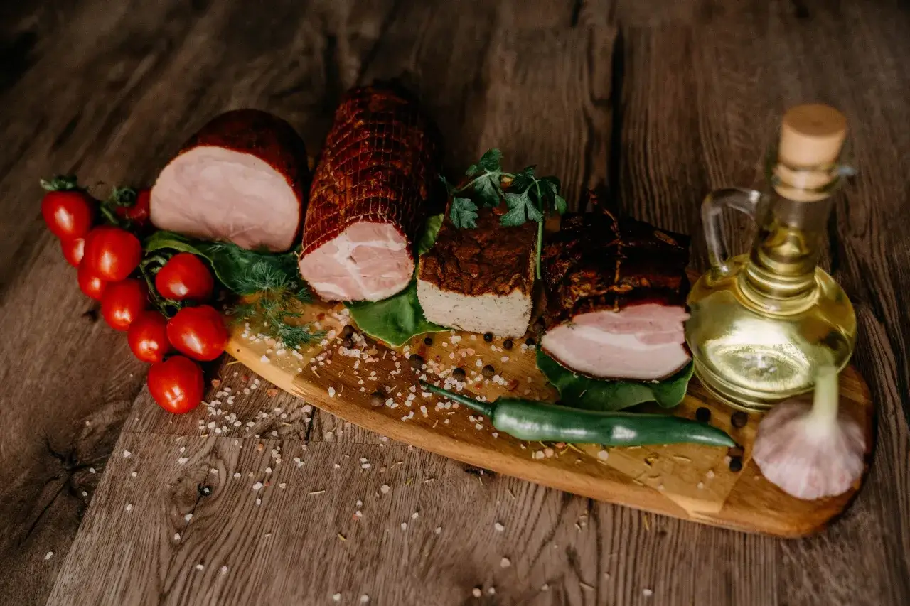 Na desce drewnianej ułożone swojskie wyroby wędliniarskie: wędzonki, pasztet, pomidorki, papryczka chili, czosnek i oliwa.