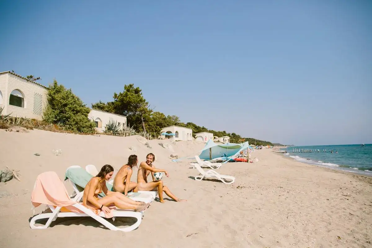 Trzy osoby opalają się na leżakach na plaży. W tle widać domki kempingowe i morze. FKK camping w pełnym słońcu.