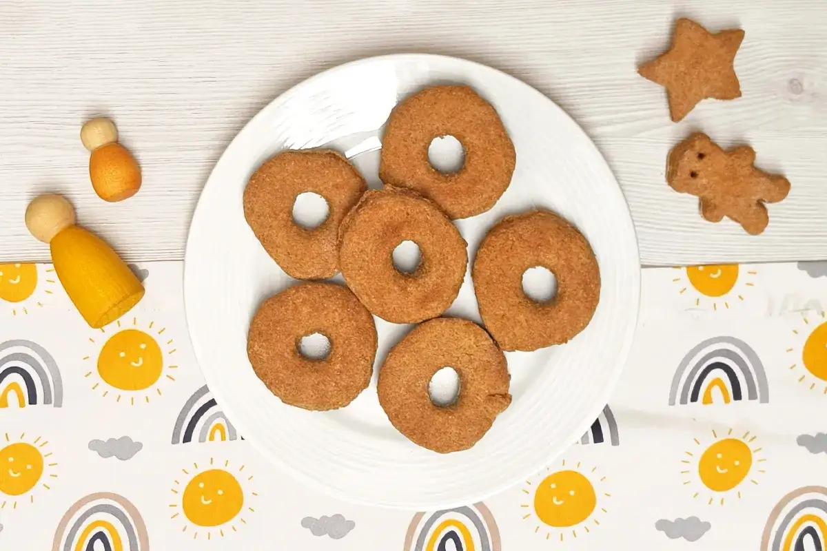 Galletas de espelta en forma de rosquilla, estrella y oso, junto a figuras de madera y un mantel con soles y arco&iacute;ris.