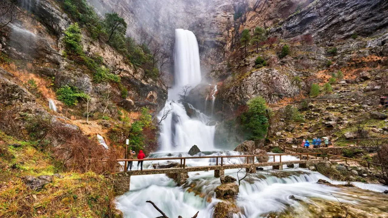 Impresionante cascada y río en un paisaje rocoso, ideal para senderismo en Castilla La Mancha. Personas observan la vista desde un puente.