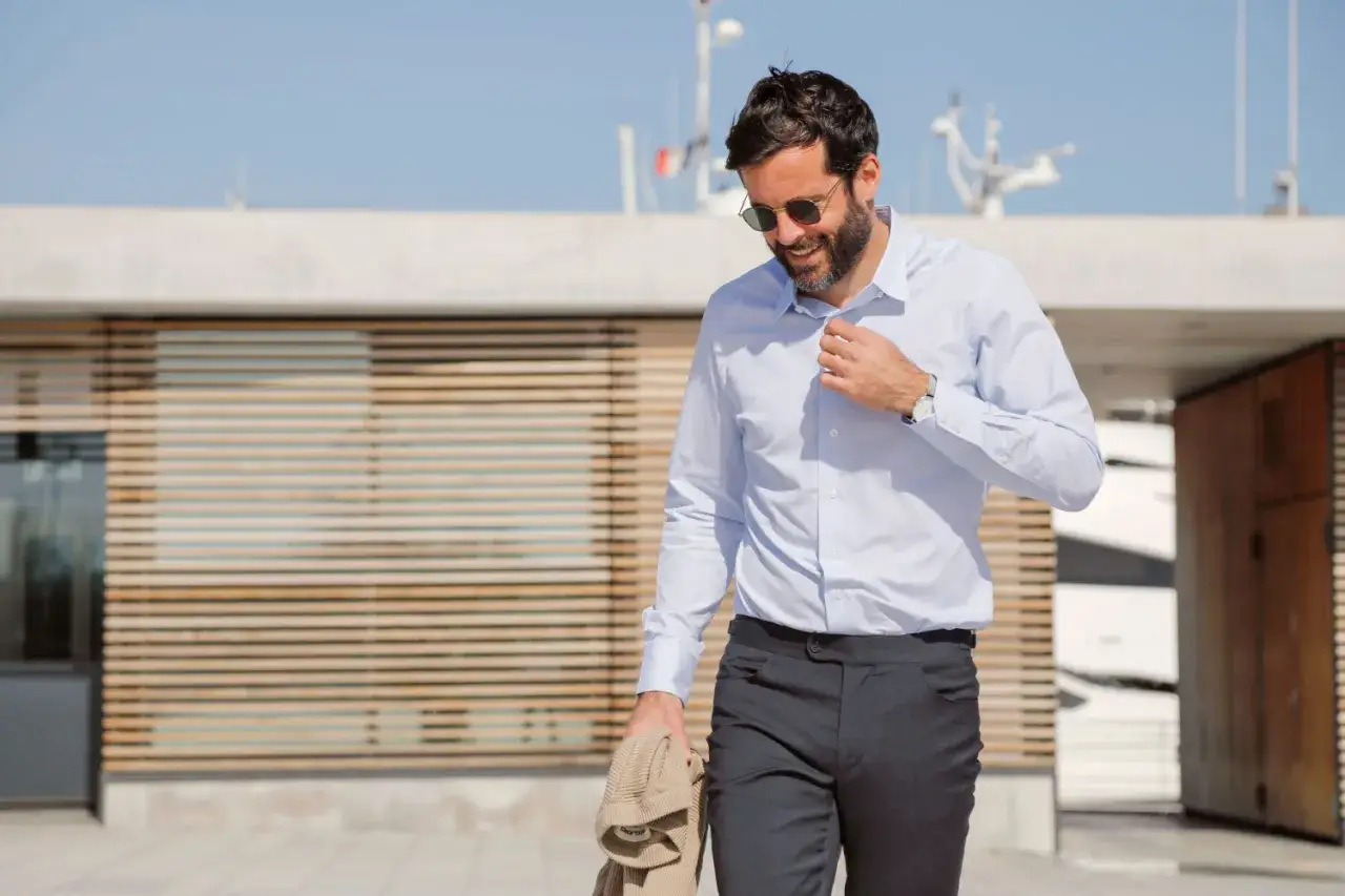 Homme souriant portant un pantalon costume sans ceinture, chemise bleue et lunettes de soleil, tenant un pull beige.