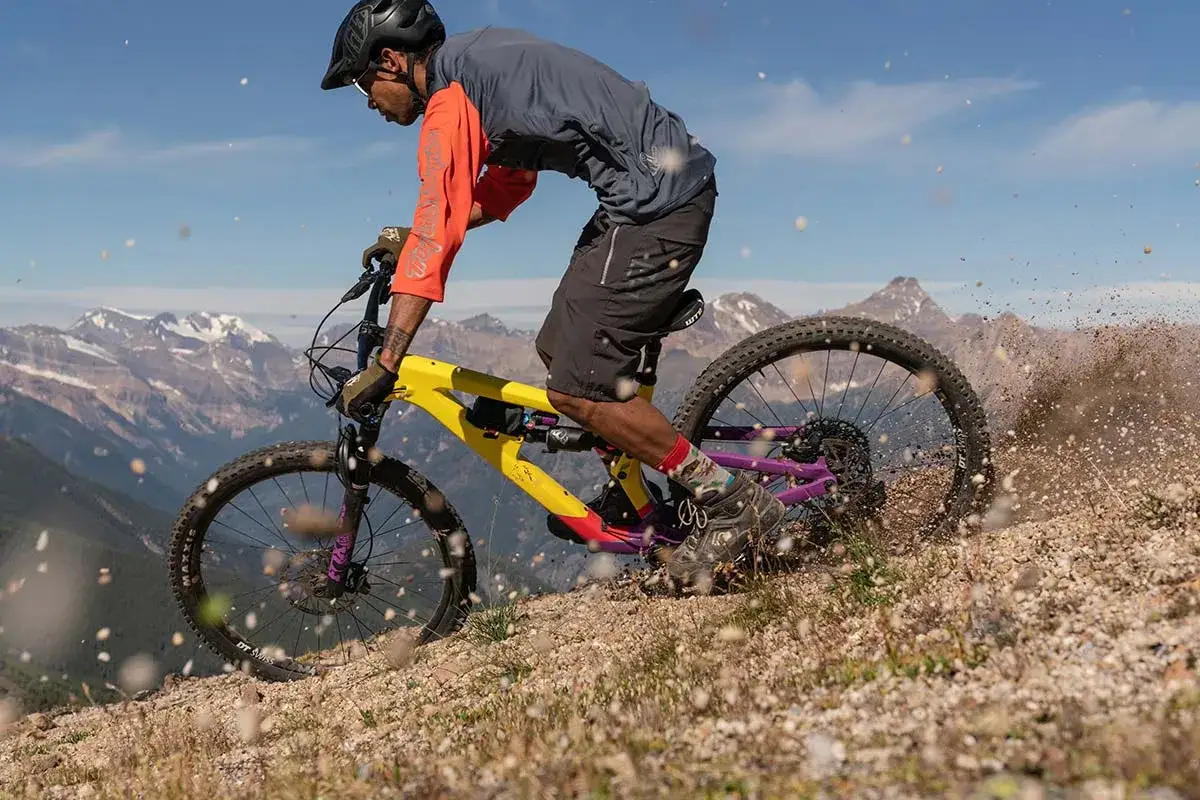 A mountain biker on a yellow and purple Salsa Cassidy bike kicks up dirt on a rocky trail with snowy mountains in the background.